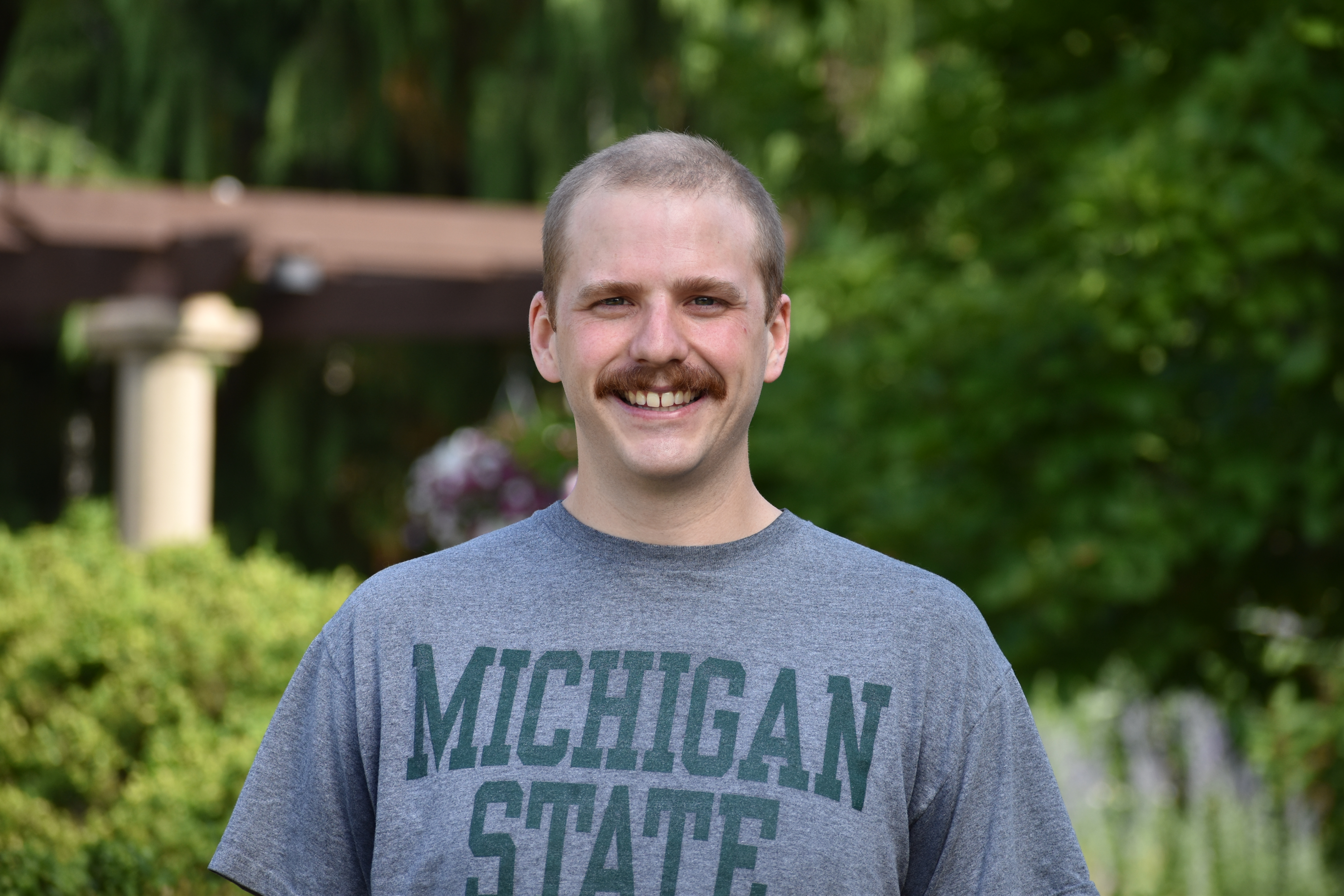 A Picture of a handsome man with short hair and a delightful mustache wearing a heather grey with green print MSU shirt